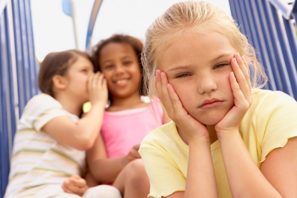 An isolated little girl with ASD looks hurt as 2 other girls tease her behind her back.