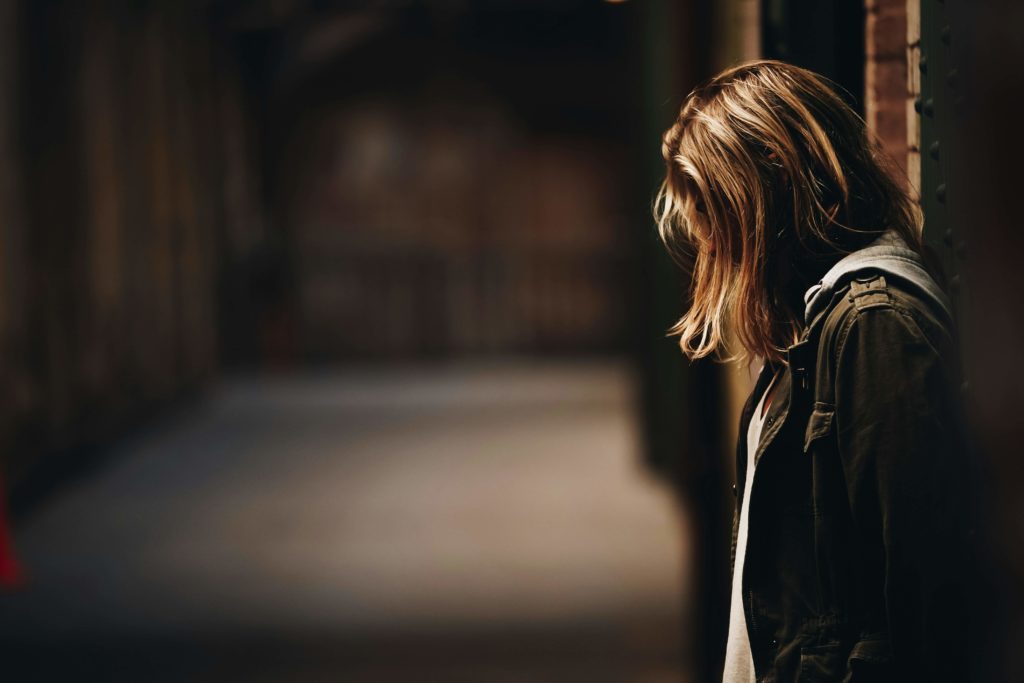 A depressed woman wearing a coat leaning against a wall and looking at her feet.