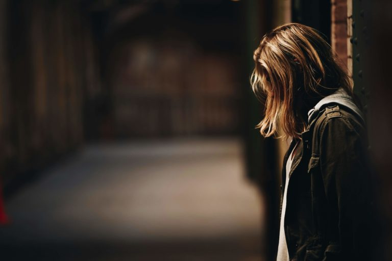 A depressed woman wearing a coat leaning against a wall and looking at her feet.