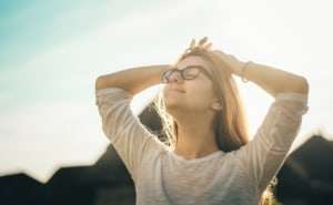 A stress-free woman in glasses standing in front of a house, looking up with eyes closed and hands behind her head.