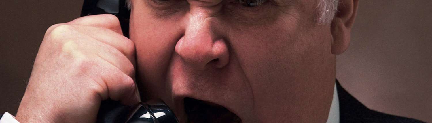 An angry and belligerent older man wearing a suit is yelling into a telephone with an enraged expression.