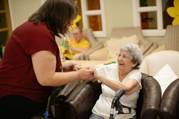 A caregiver helping a smiling elderly lady stand up from a chair.