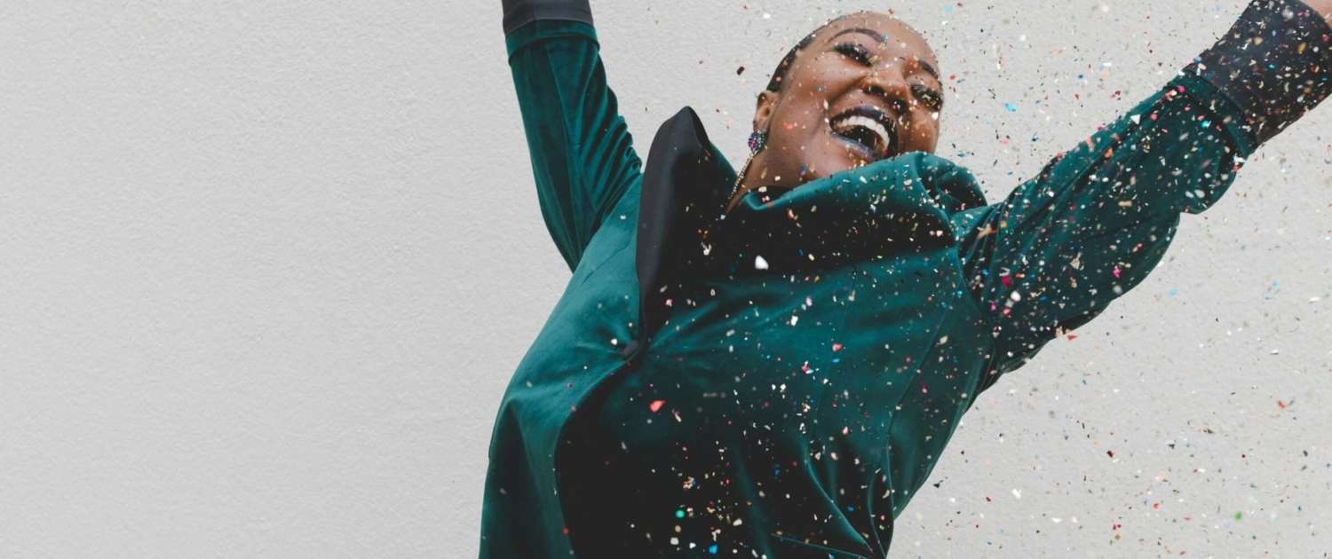 African American woman jumping with joy and throwing confetti in celebration. Celebrate yourself.