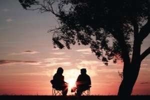 A couple sitting in lawn chairs at sunset talking next to a silhouetted tree.