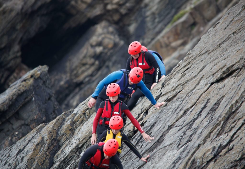 Five people wearing climbing gear show trust as they follow their guide along a steep rock cliff.