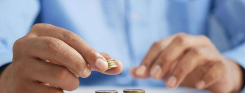 A man in a blue shirt is shown stacking coins in equal piles. Budgeting is the number one reason for conflict in couples.