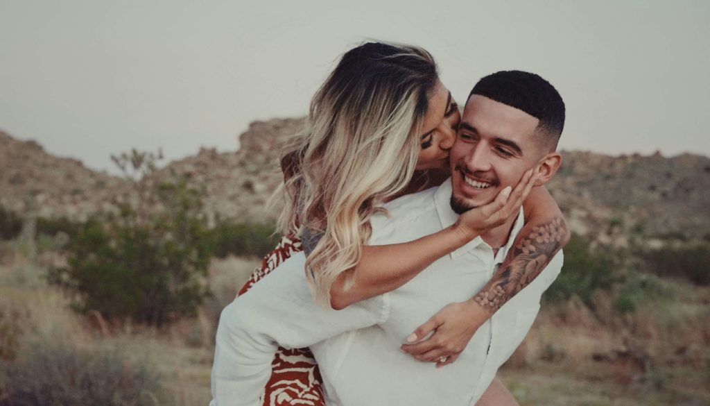 A woman riding piggyback on a man, kissing him as he smiles.