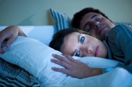 Image of a lady awake in bed with her husband sleeping peacefully behind her