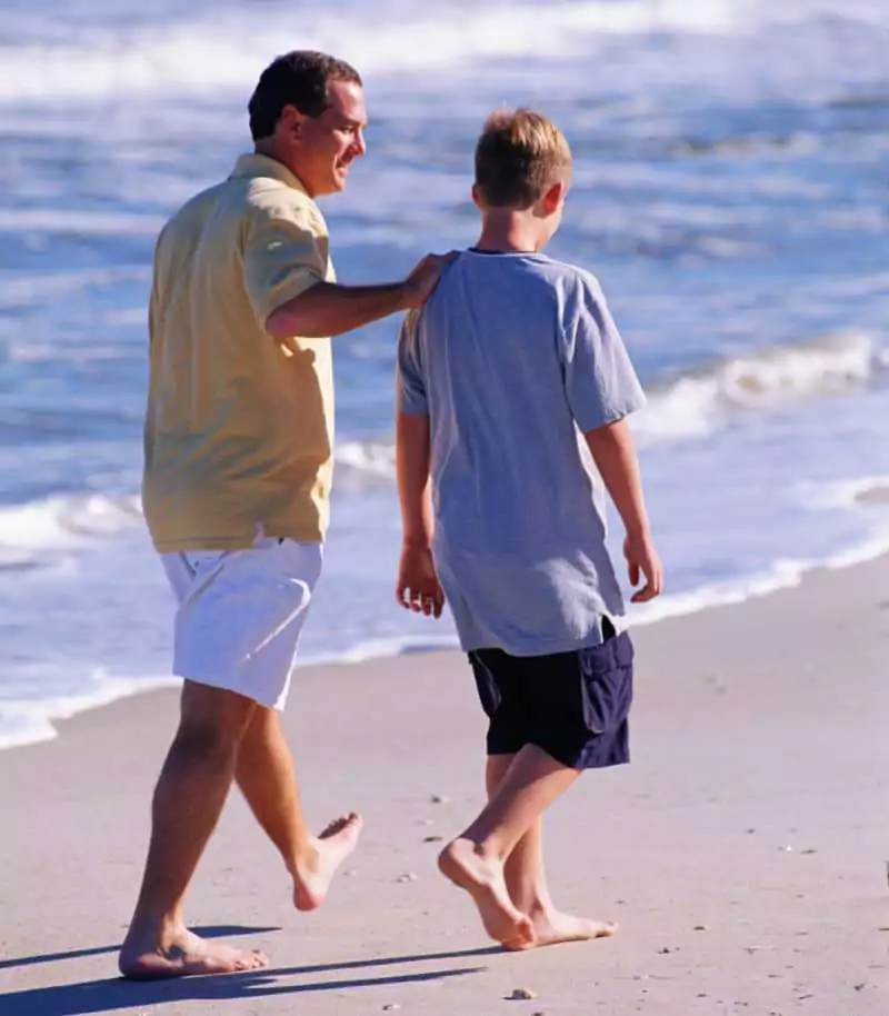 Father and son walking on a beach with father touching son's shoulder.