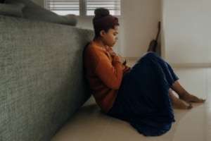 A woman having a panic attack holds her chest while sitting on the floor leaned against a sofa.