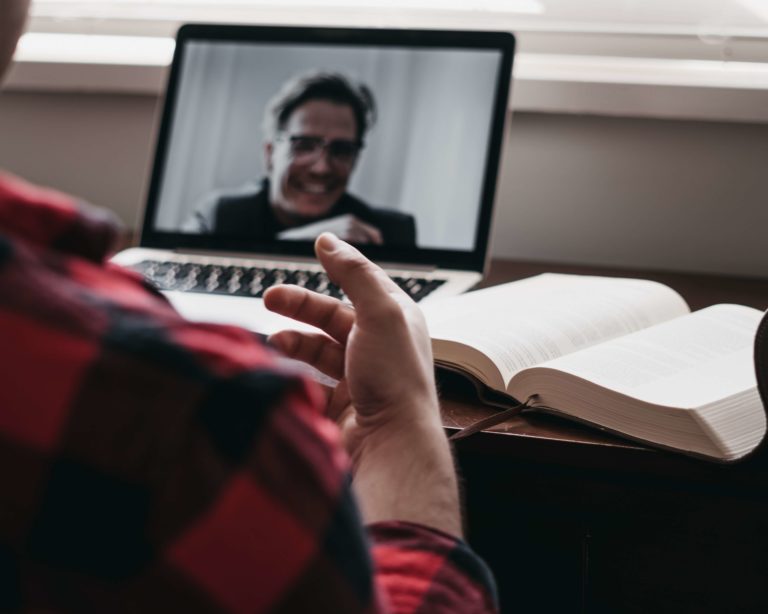 Looking over the shoulder of a man in a red and black shirt who is looking at a counselor on the screen of his laptop.