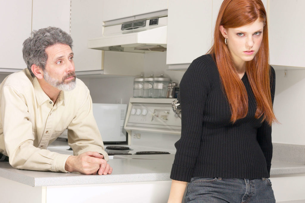 Father leaning on kitchen counter talking to teen girl who has her back turned to him.