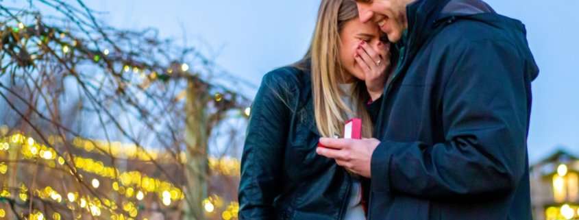 A happy and surprised woman is snuggled against her boyfriend who is holding an engagement ring