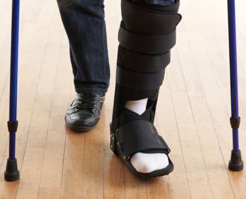 An image of a person's lower legs and feet, with a support boot on their left foot. There are 2 blue crutches outside of their legs.