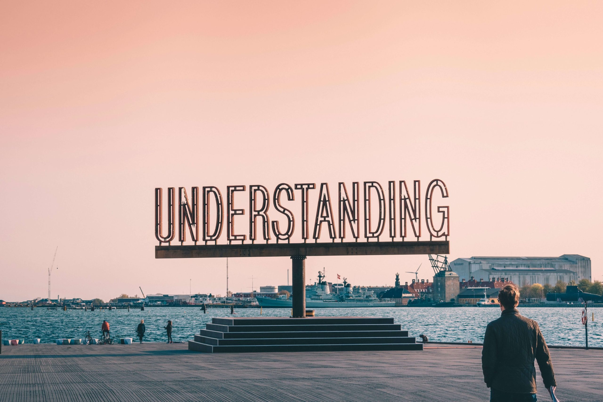 Large "Understanding" sign sculpture by the water at sunset, with a person in the foreground.