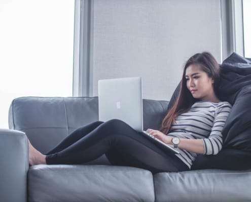 Asian woman wearing a striped shirt reclining on a sofa with a laptop during a video counseling session.